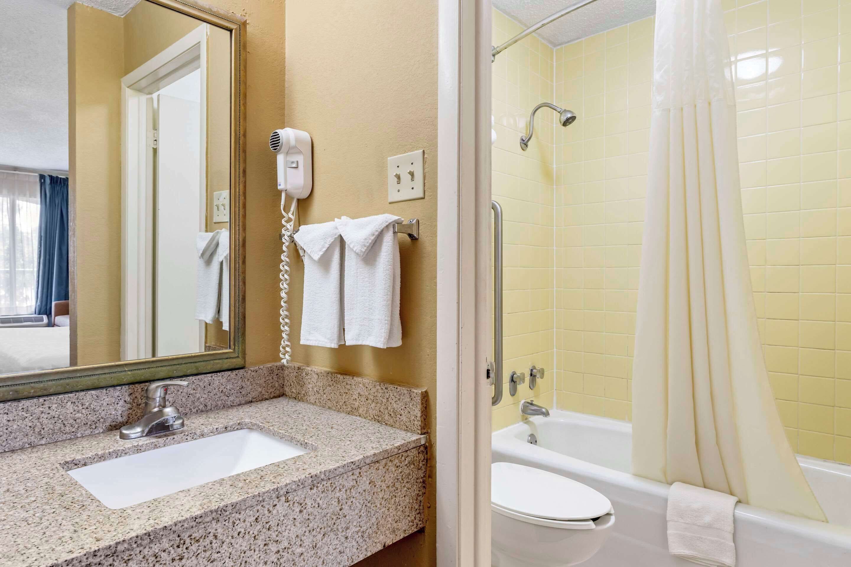 Une salle de bain avec un lavabo surmonté d’un plan de travail en granit, un miroir, un sèche-cheveux, une douche aux carreaux jaunes et une baignoire blanche.