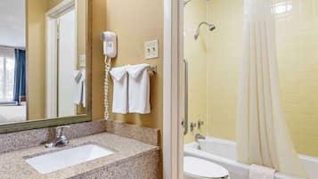 Une salle de bain avec un lavabo surmonté d’un plan de travail en granit, un miroir, un sèche-cheveux, une douche aux carreaux jaunes et une baignoire blanche.