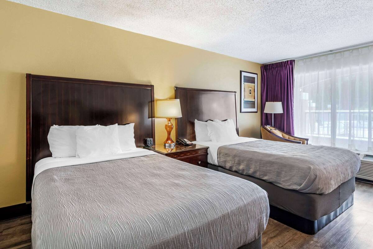 A hotel room with two beds, a nightstand with a lamp, a telephone, and a window with curtains.