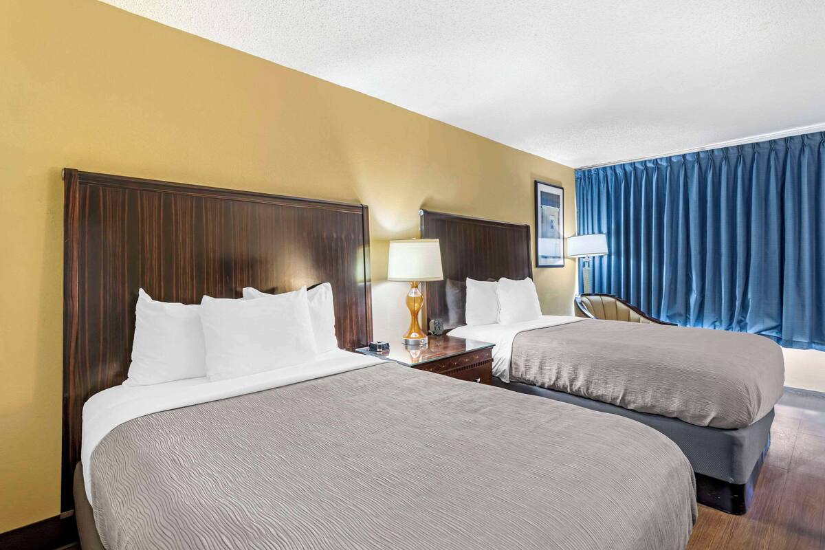 A hotel room with two beds, a nightstand with a lamp, a chair, and a large window with curtains.