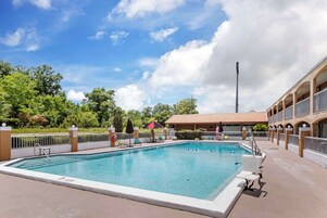 Una piscina rectangular con borde blanco, rodeada por una valla baja. Hay sillas de descanso y sombrillas cerca de la piscina, y un edificio con techo de tejas al fondo.