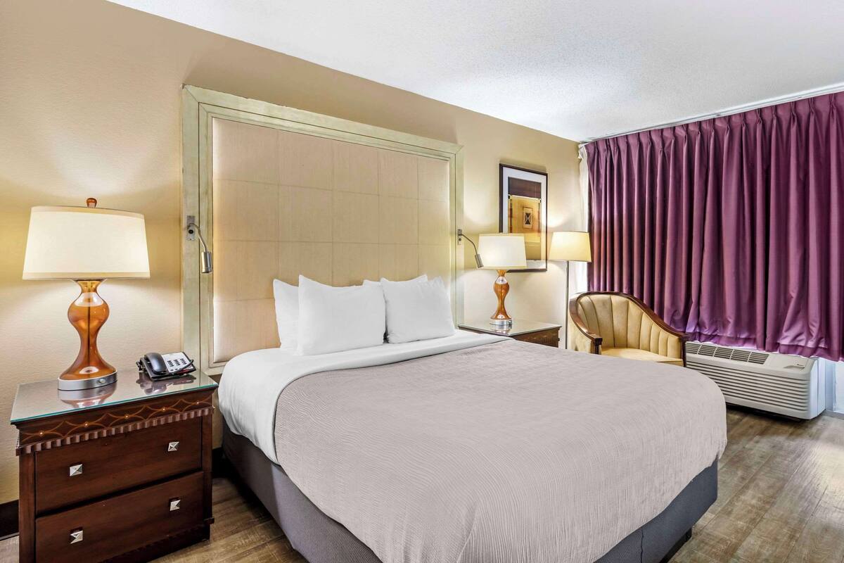 A hotel room with a large bed, two bedside tables with lamps, a chair, a wardrobe, and a window with curtains.