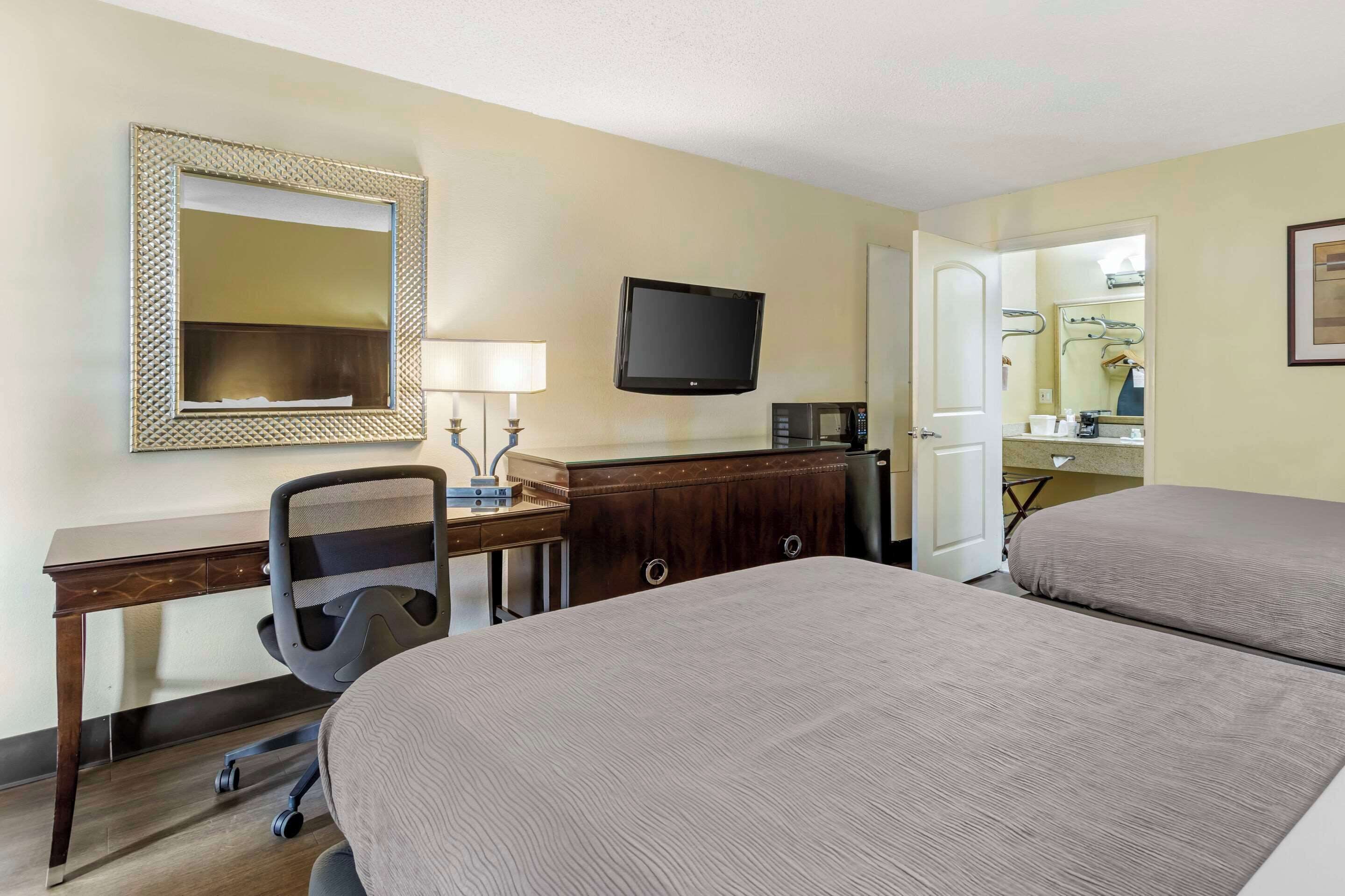 A hotel room with two beds, a desk with a chair, a television, a microwave, and a bathroom visible through an open door.