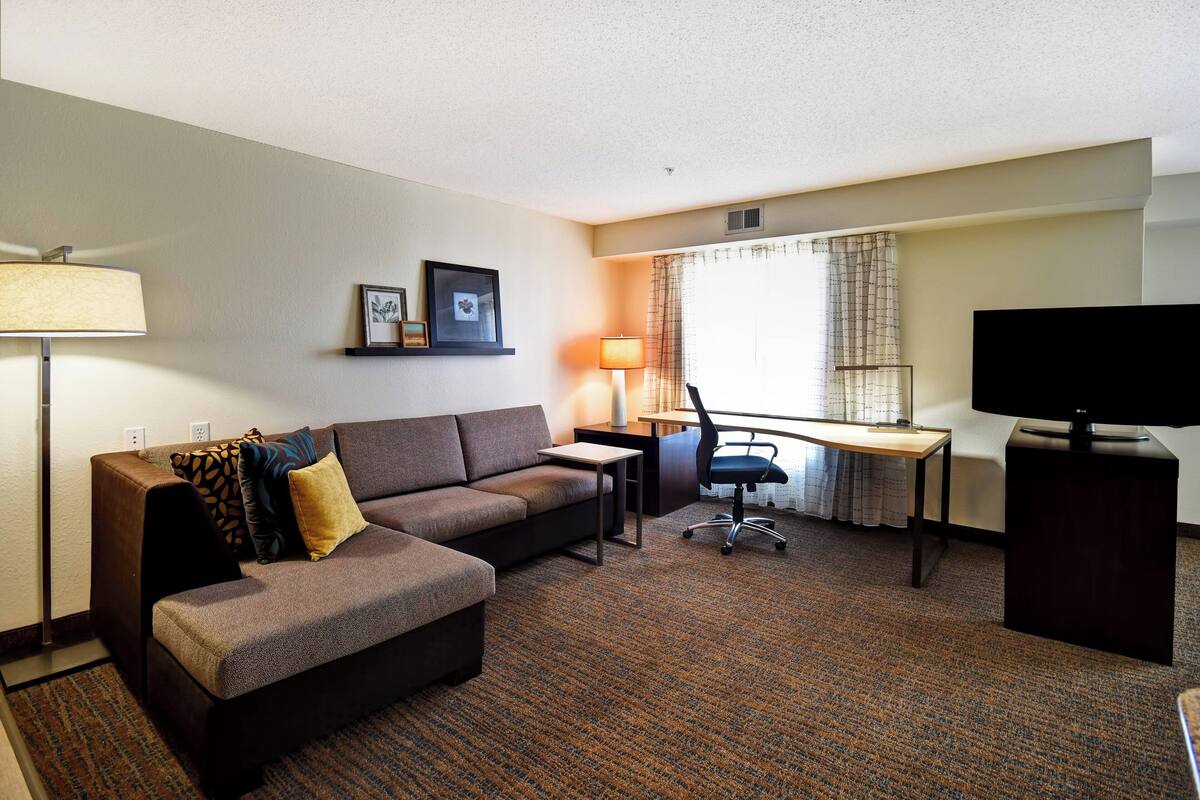 A hotel room with a sofa, desk, chair, TV, lamp, and a window with curtains.