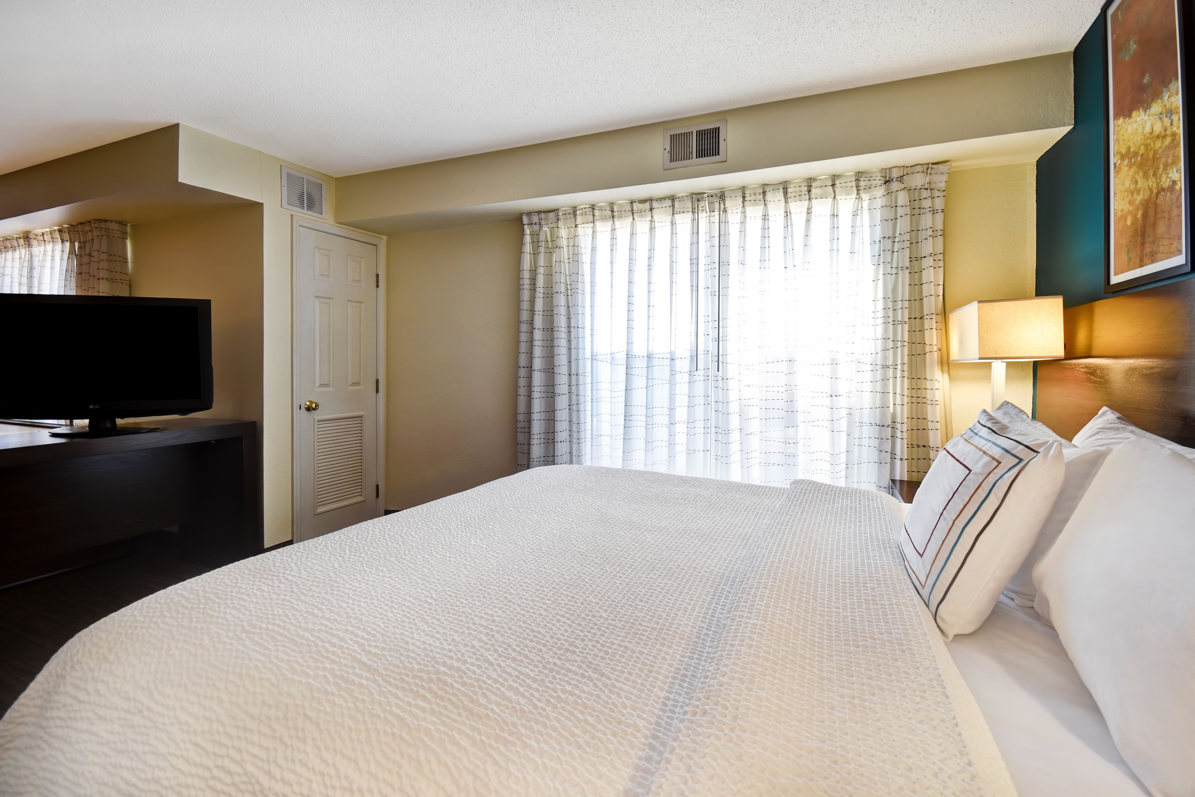 Habitación de hotel con una cama grande, un televisor sobre un soporte, una puerta y una ventana con cortinas.