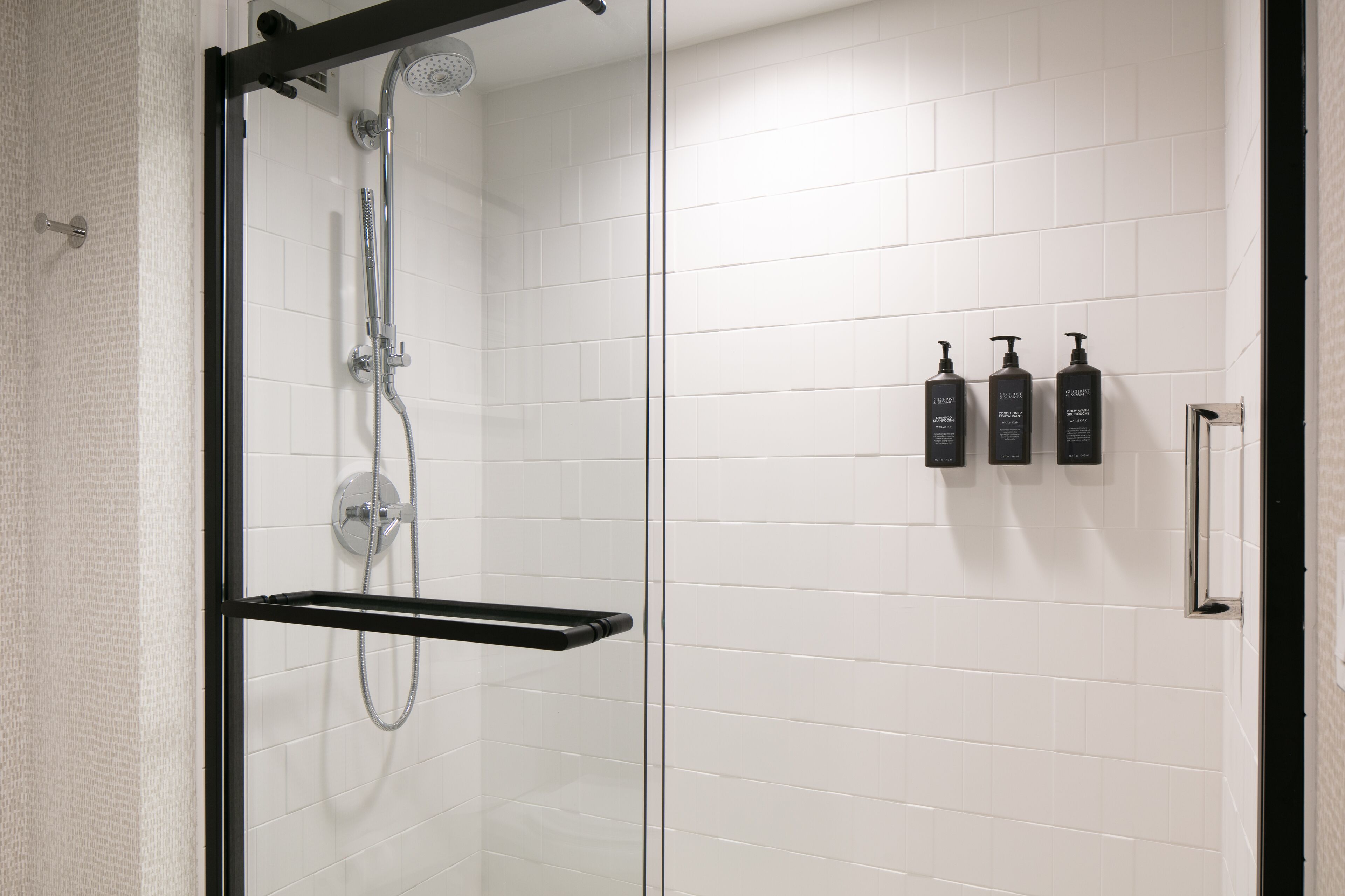 Une salle de bain moderne avec une douche à parois de verre, des murs recouverts de carreaux blancs et trois bouteilles de shampoing noires fixées au mur.