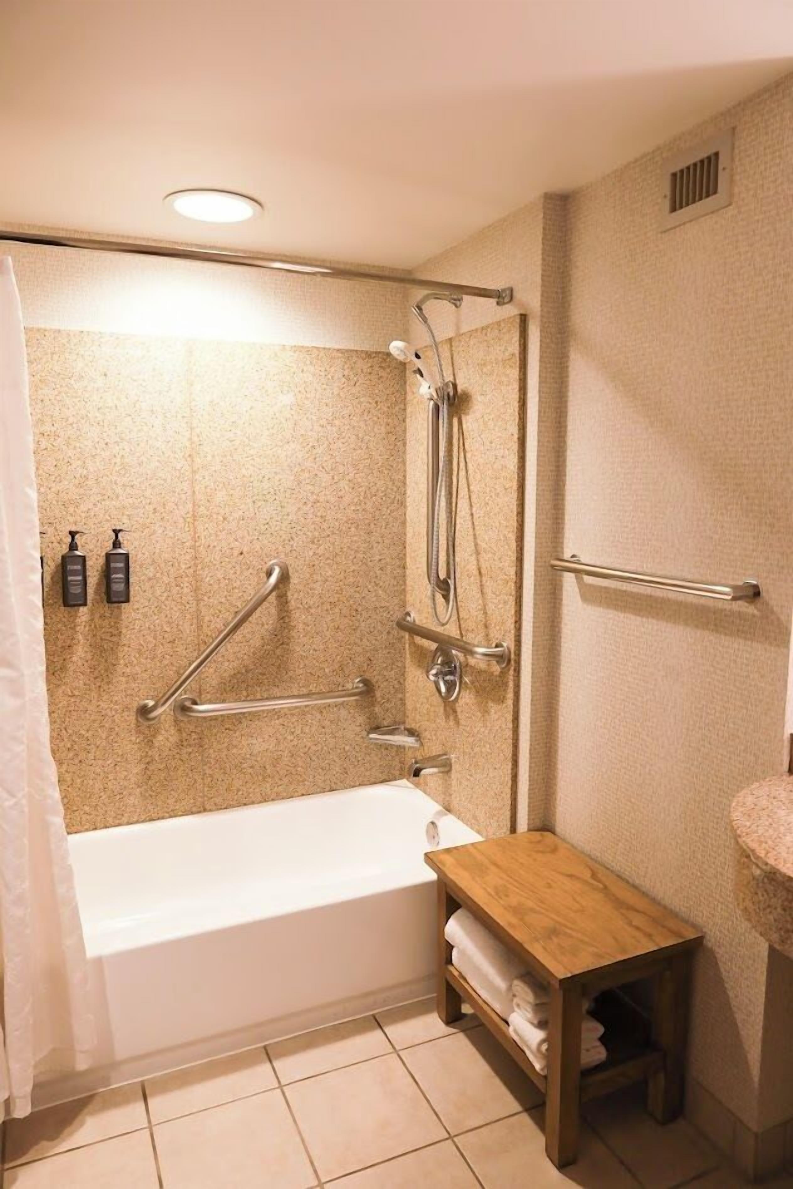 A bathroom with a bathtub, grab bars, a wooden shelf, and a towel rack.