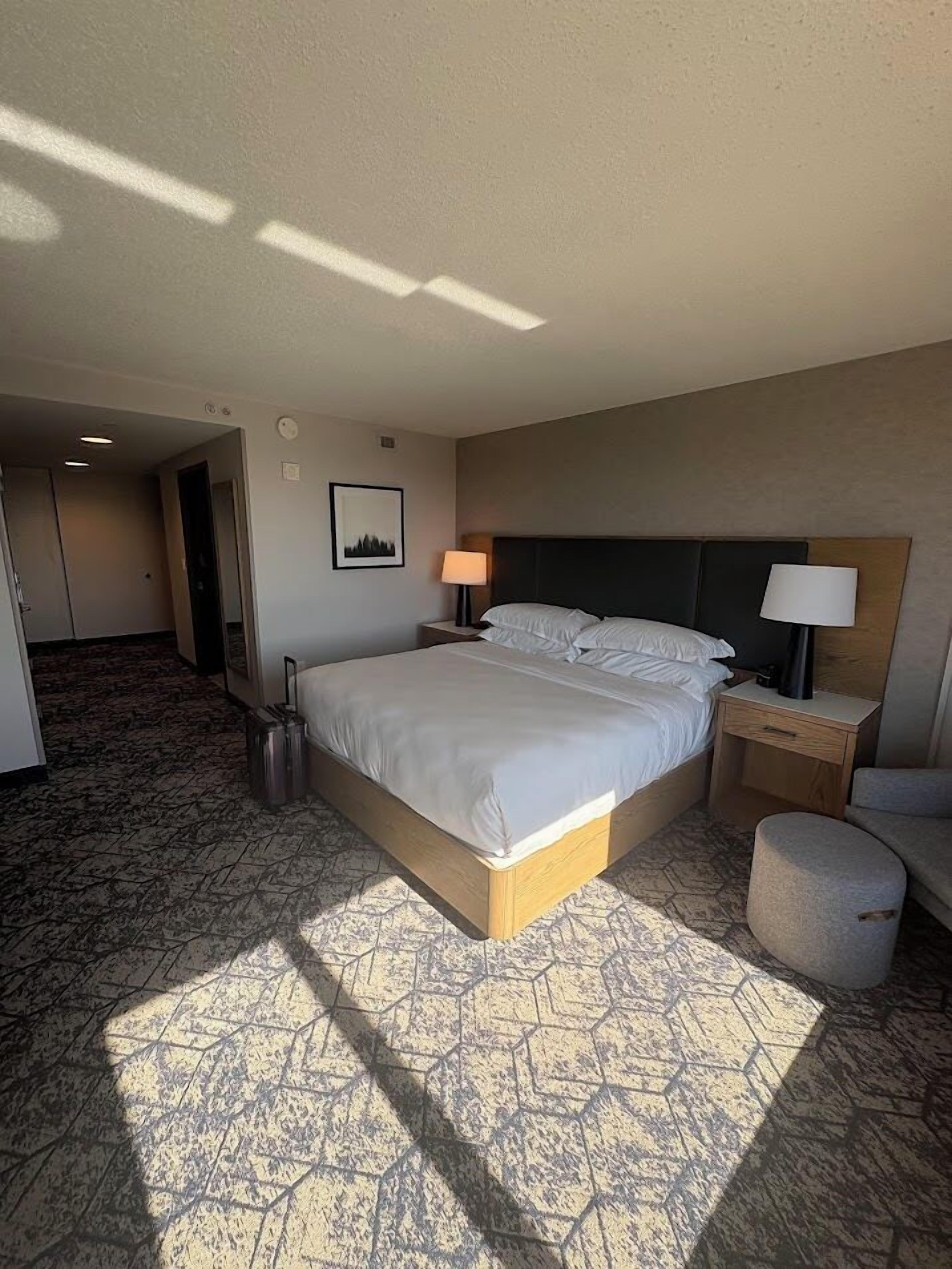 A hotel room with a large bed, two bedside tables with lamps, a small ottoman, and a carpeted floor.