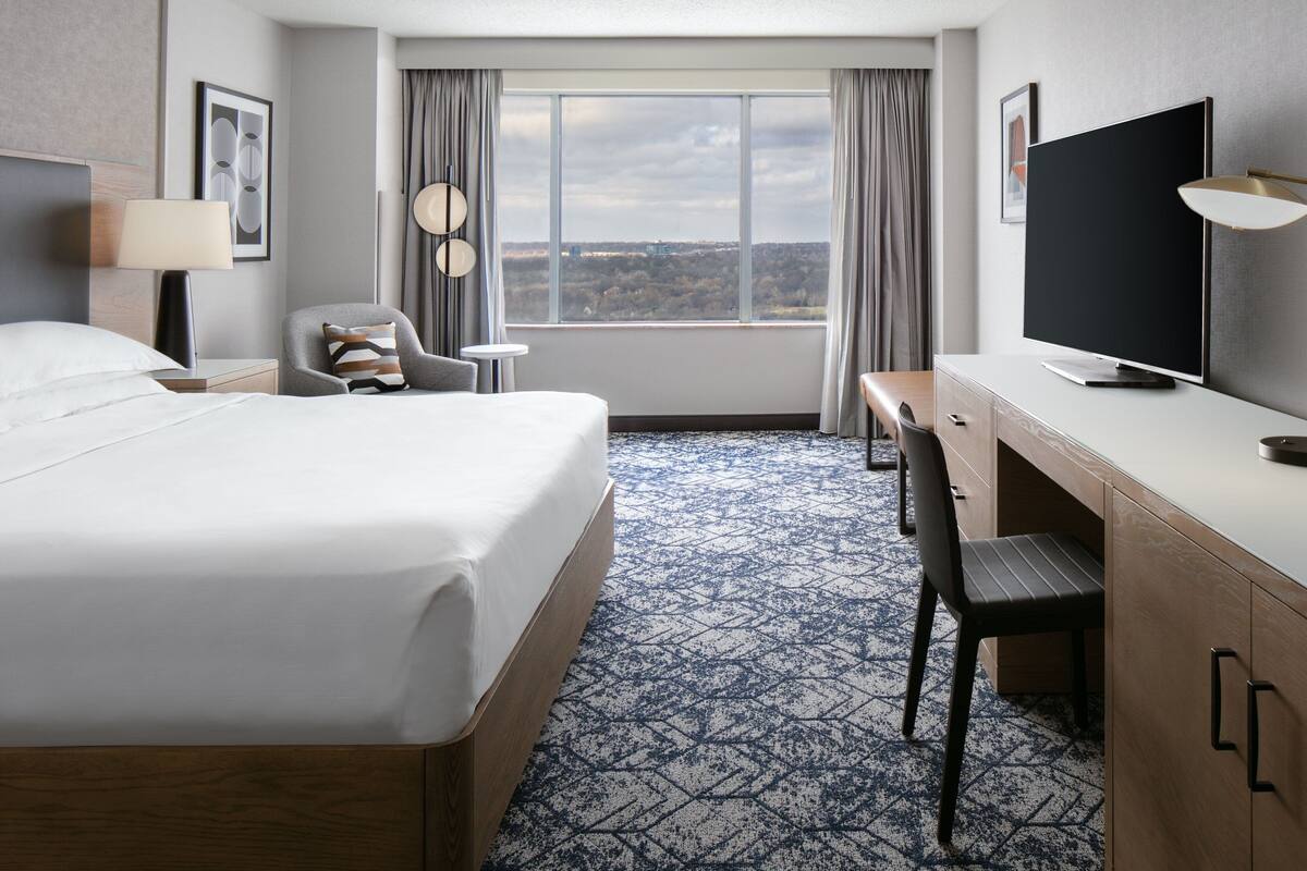 A hotel room with a large bed, a desk with a chair, a television, and a view of the city through the window.