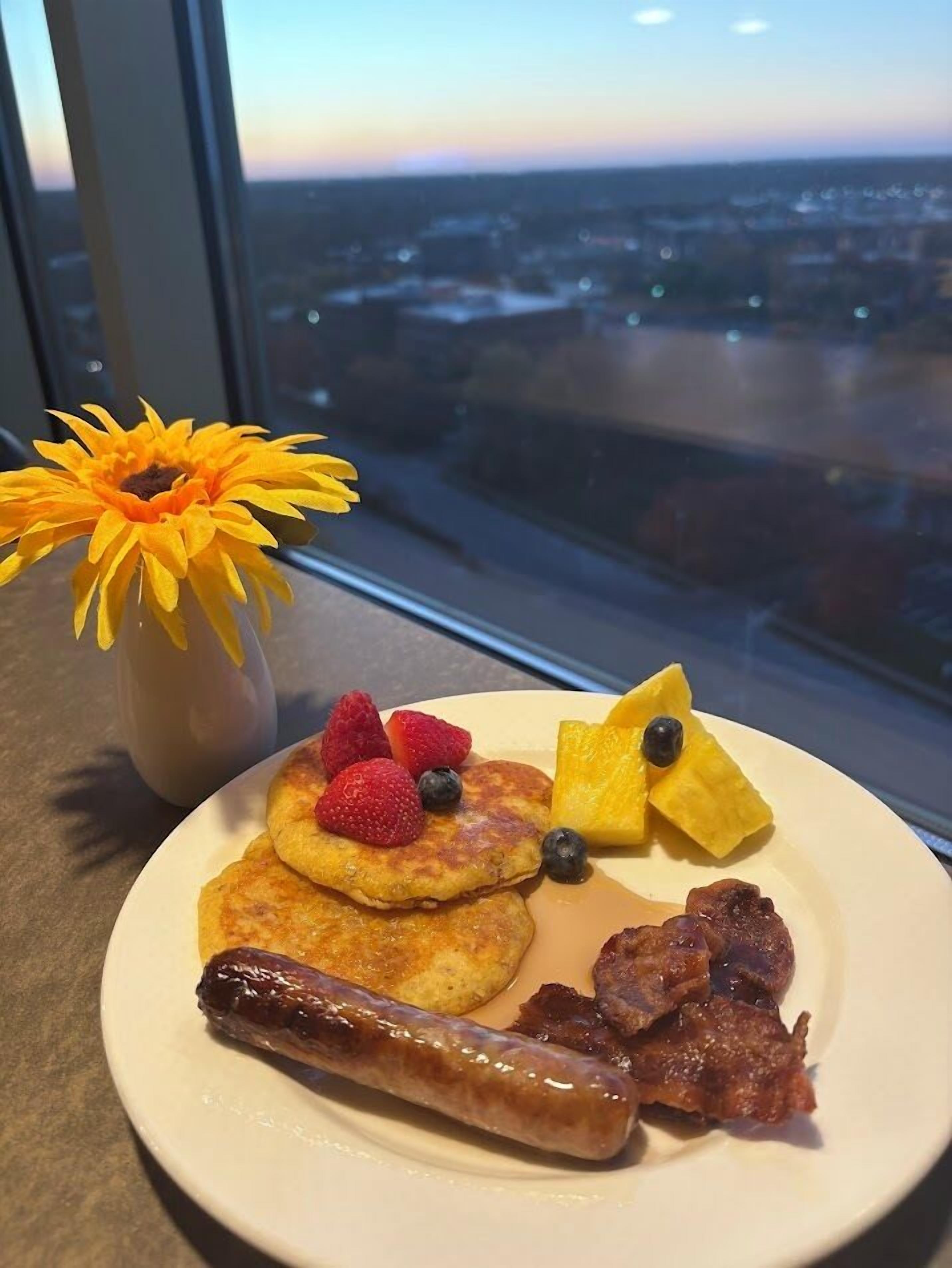 Une assiette de petit-déjeuner avec des crêpes, des fraises, des myrtilles, une saucisse et du bacon, et un vase de fleurs jaunes placé sur une table près d’une fenêtre donnant sur un paysage urbain.