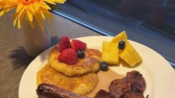 Une assiette de petit-déjeuner avec des crêpes, des fraises, des myrtilles, une saucisse et du bacon, et un vase de fleurs jaunes placé sur une table près d’une fenêtre donnant sur un paysage urbain.