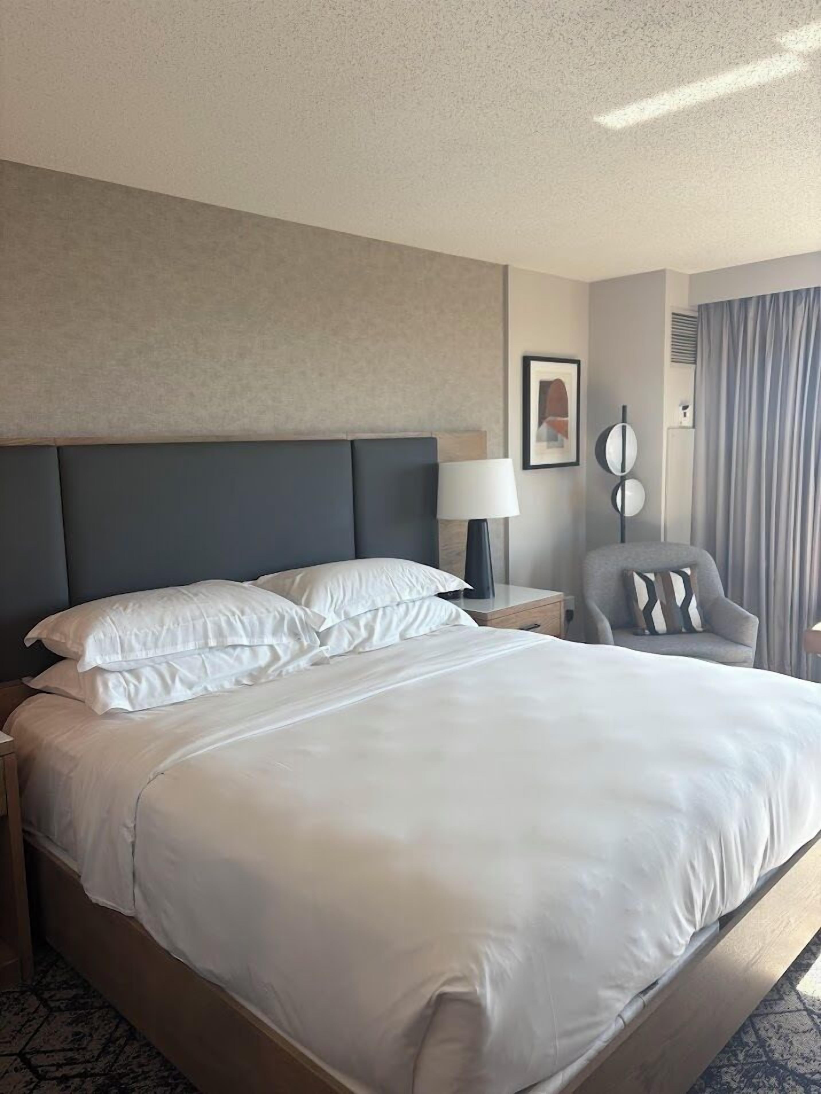 Een hotelkamer met een groot bed, een stoel, een nachtkastje, een lamp en een spiegel aan de muur.
