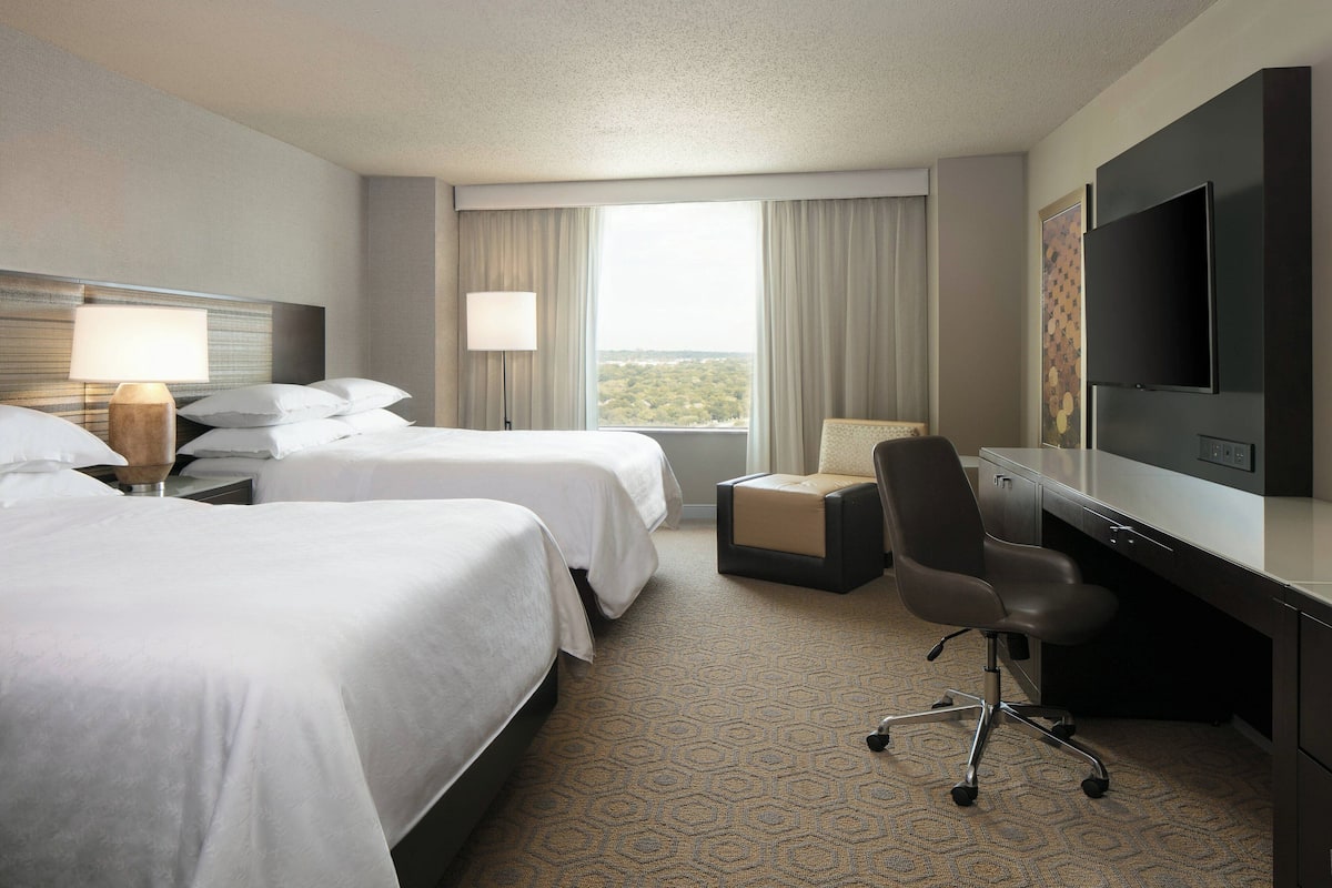 A hotel room with two beds, a desk, a chair, a TV, and a large window with a view.