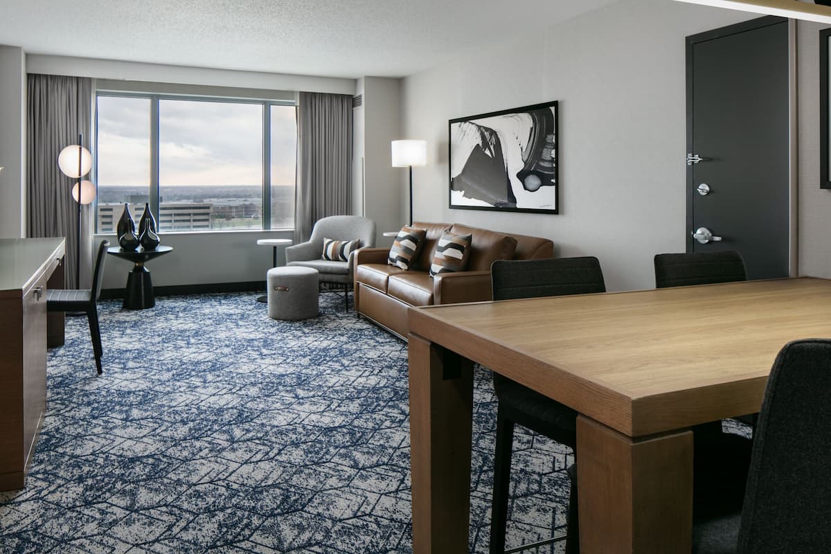 A hotel room with a large window, a sofa, a dining table, and a lamp.