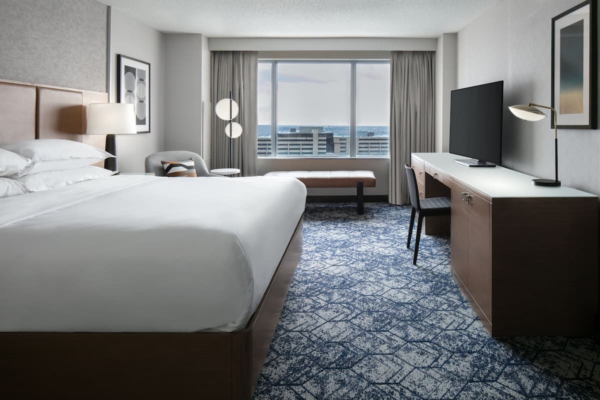 A hotel room with a large bed, a desk with a flat-screen TV, a chair, a desk lamp, and a window with a city view.