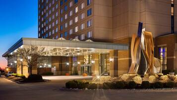 A modern hotel entrance with a glass facade, a large sculpture, and a parking area.