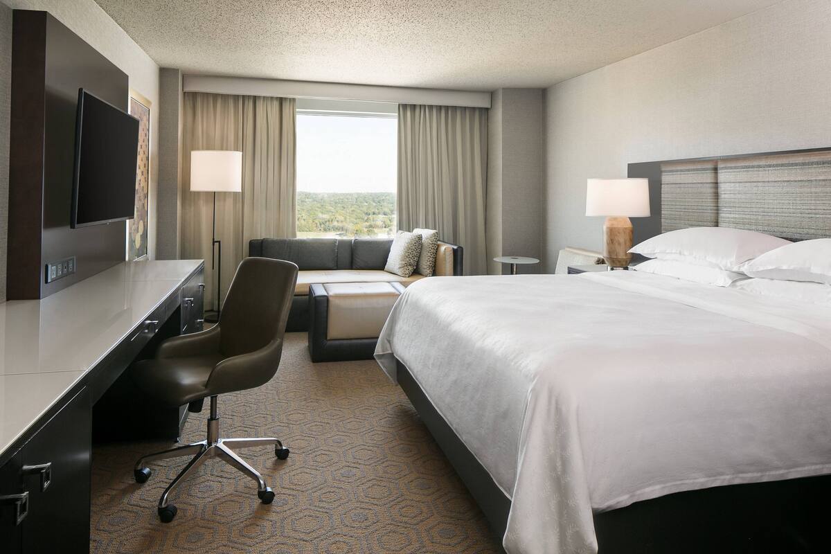 A hotel room with a bed, a desk, a chair, a TV, and a window with curtains.