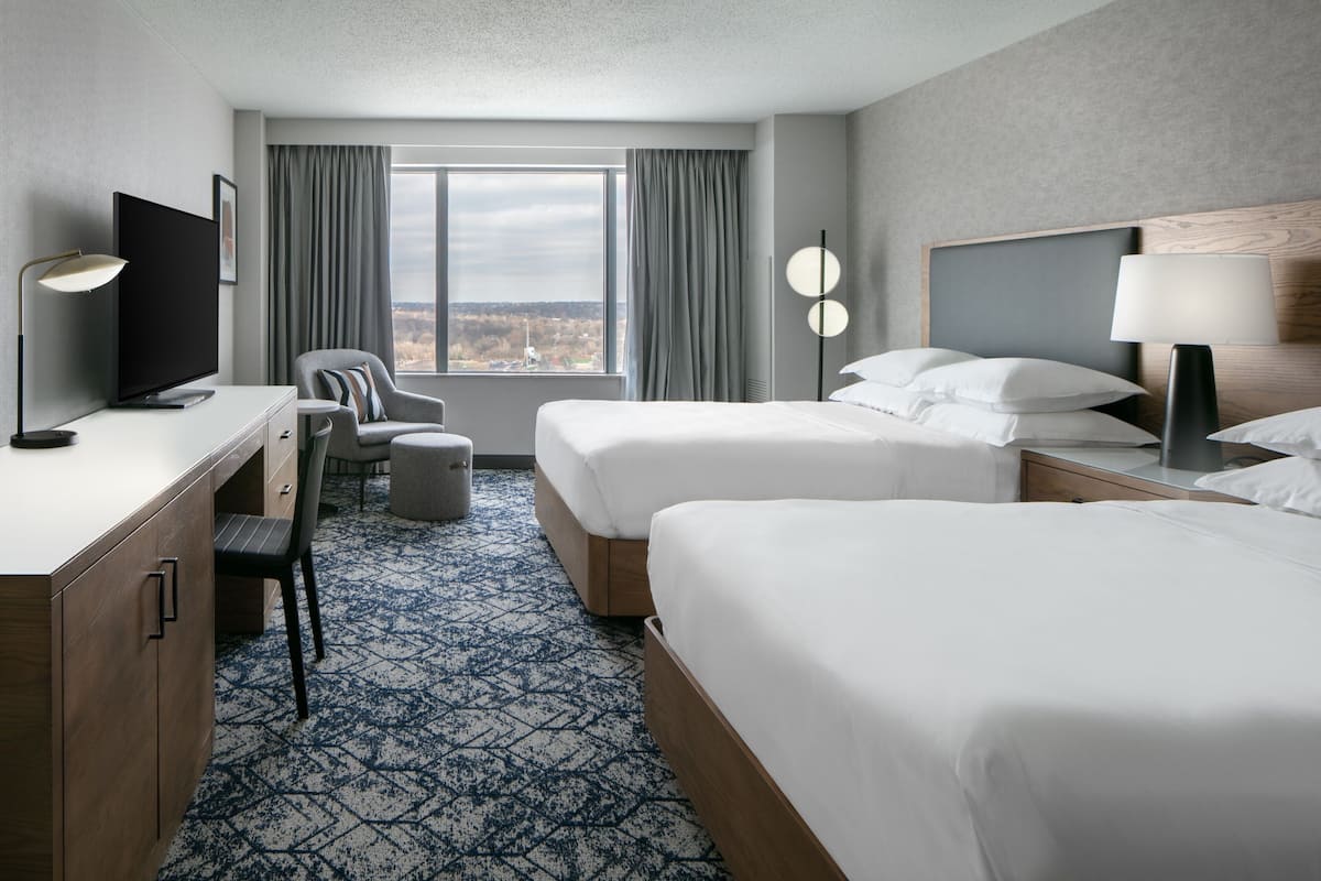 A hotel room with two beds, a desk, a chair, a television, and a view of the city through the window.