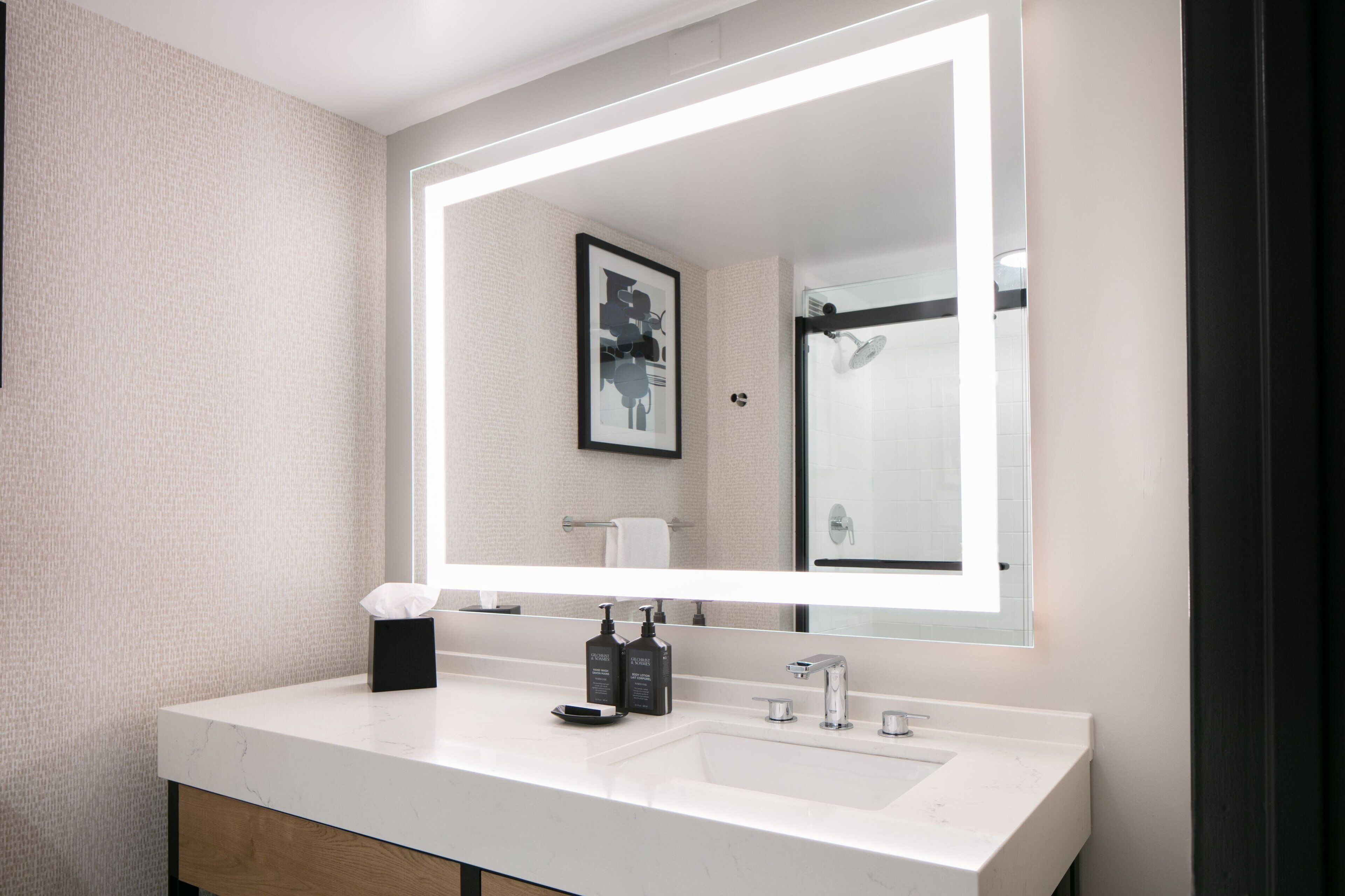 A modern bathroom with a large mirror, white marble countertop, and a black soap dispenser.