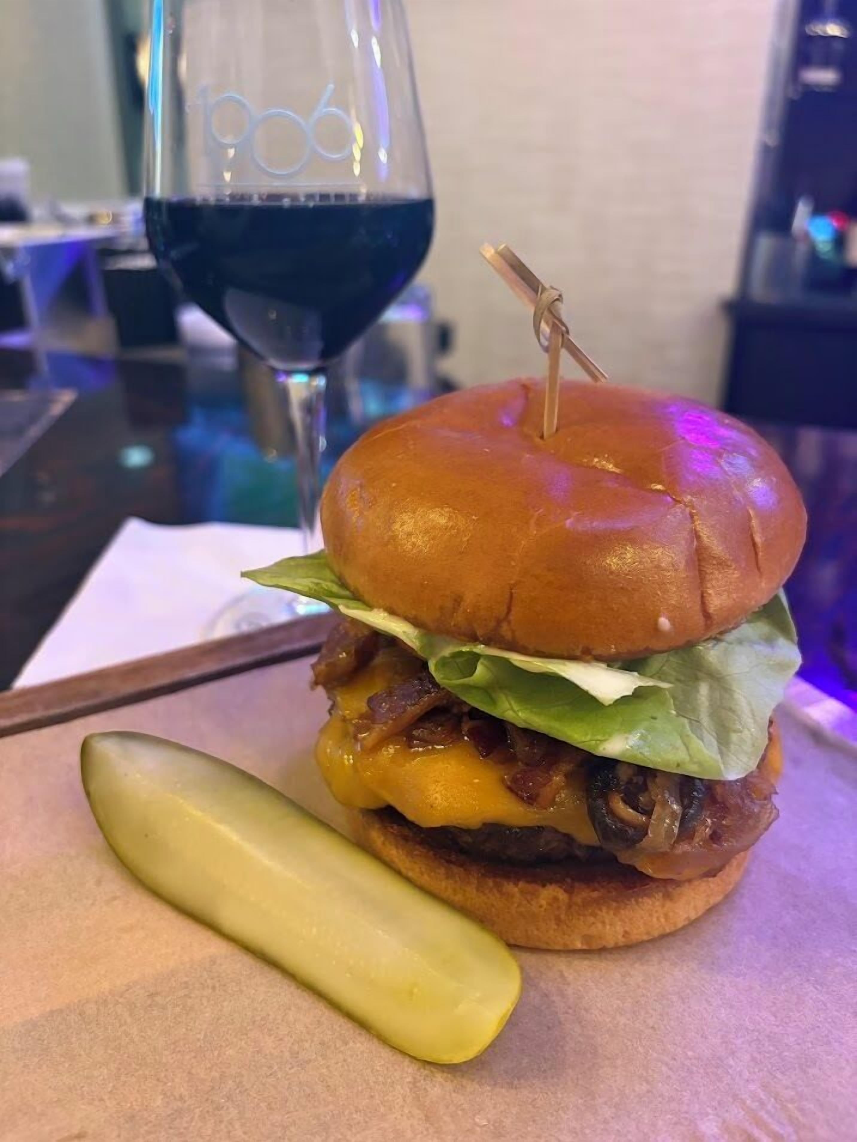 Un hamburger accompagné d’un cornichon, un verre de vin rouge et un bar en arrière-plan.