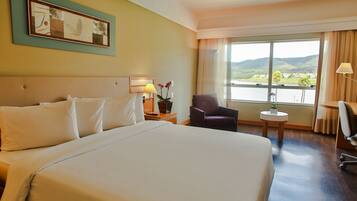 Habitación de hotel con una cama grande, una silla, una mesita, una lámpara y una ventana con vistas a las colinas.