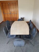 Un salón de conferencias con una mesa rectangular, ocho sillas tapizadas, un cesto de basura pequeño y una puerta de madera.