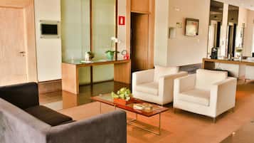 A modern hotel lobby with a reception desk, two white armchairs, a coffee table with a plant, and a large mirror.