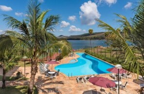 A swimming pool with a winding design, surrounded by palm trees and lounge chairs, with a view of a lake and hills in the background. - Transamerica Lagoa dos Ingleses (Nova Lima)