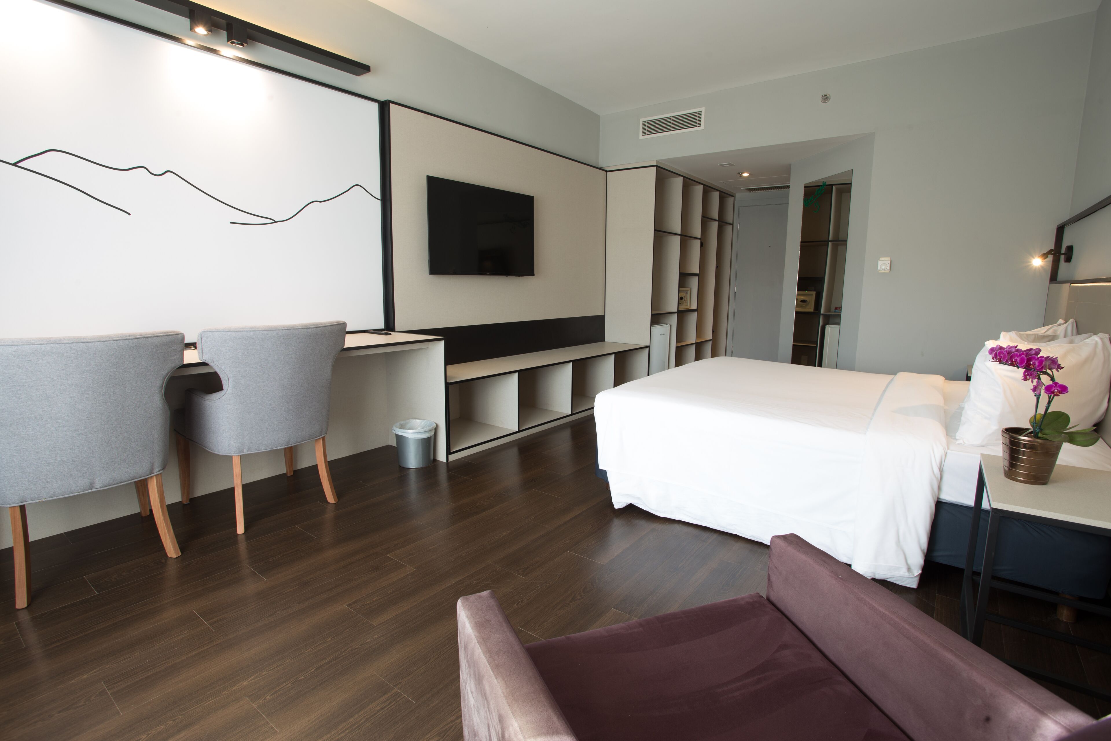 Habitació d'hotel moderna amb un llit gran, un escriptori, dues cadires, una prestatgeria i un televisor penjat a la paret.