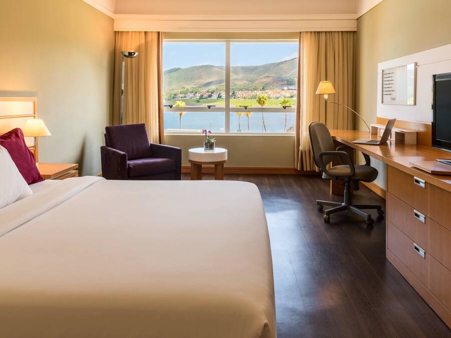 Habitación de hotel con una cama grande, un escritorio con computadora, una silla, una mesita con un florero y vistas a un lago y montañas.