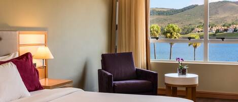 A hotel room with a bed, a chair, a nightstand with a lamp, a small table with a plant, and a large window offering a view of hills and water.