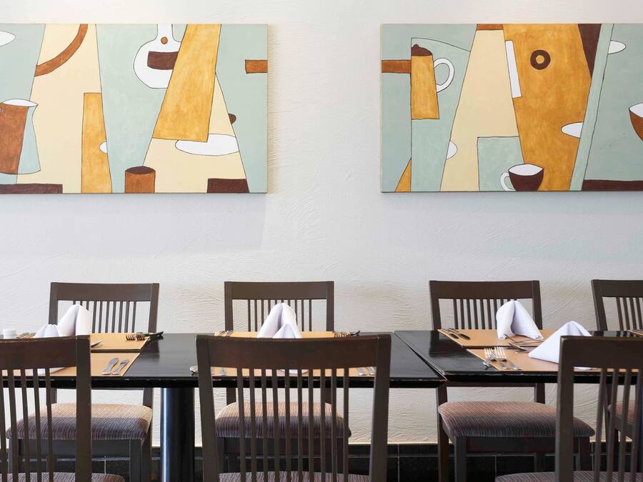 Uma área de refeições com quatro mesas e cadeiras, cada conjunto com talheres e guardanapos. Duas pinturas abstratas estão penduradas na parede acima das mesas.