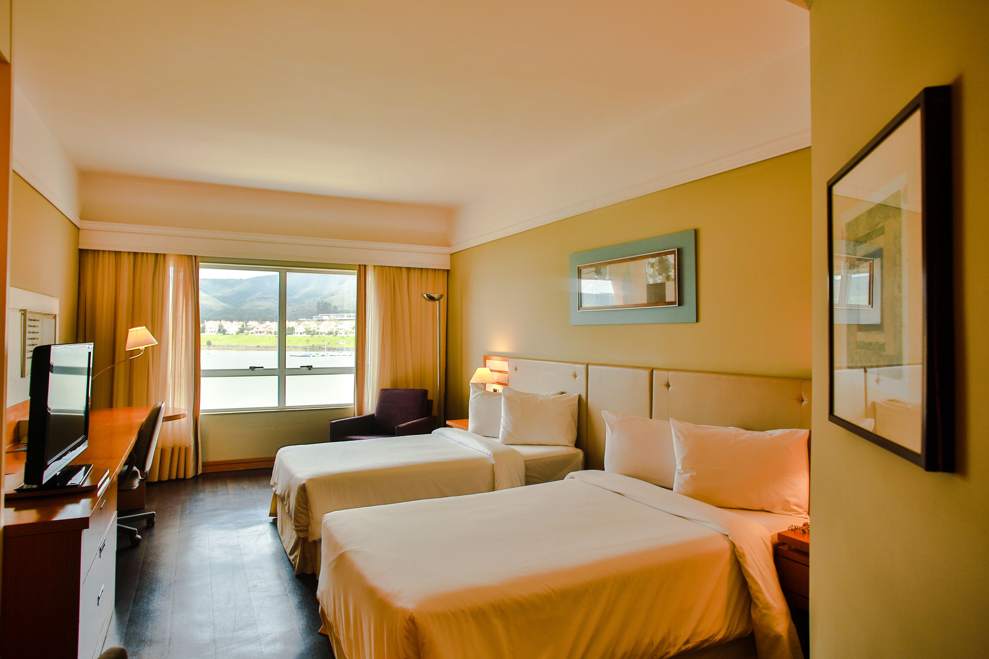 A hotel room with two beds, a desk with a TV, and a view of the outdoors.