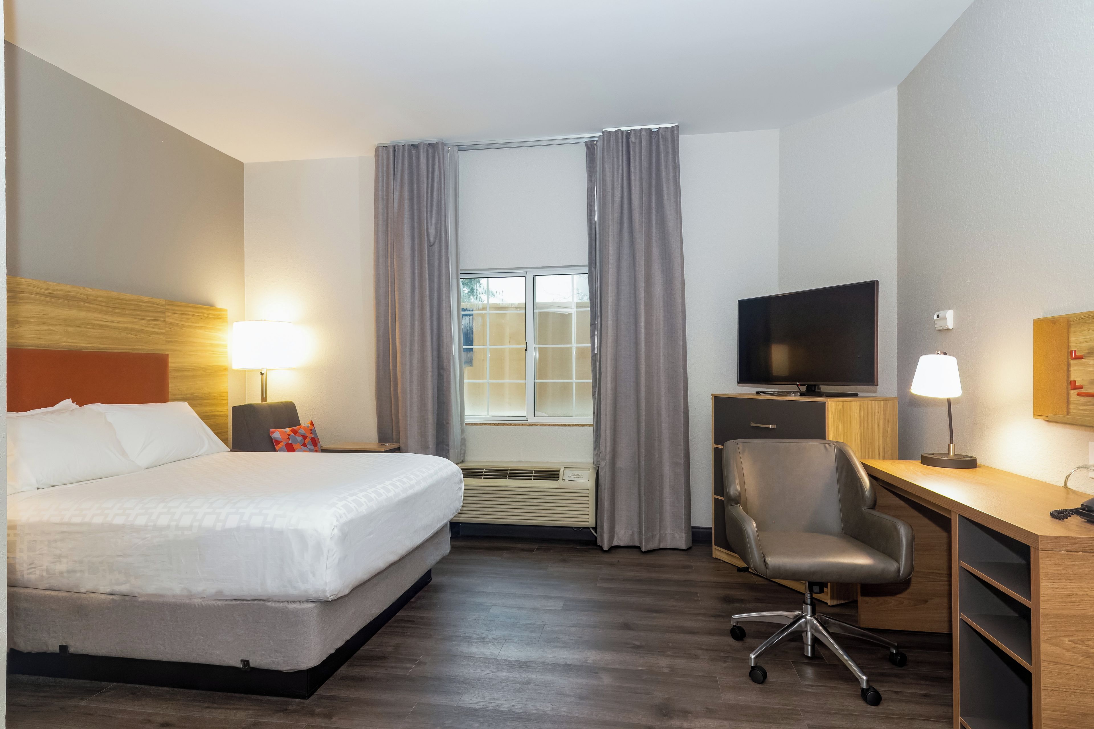 Habitación de hotel con cama, escritorio, silla, televisor y ventana con cortinas.