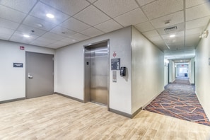 Een hotelgang met een lift, een deur met de tekst 'Elektrische Ruimte' en een kamerbreed tapijt met dessin.