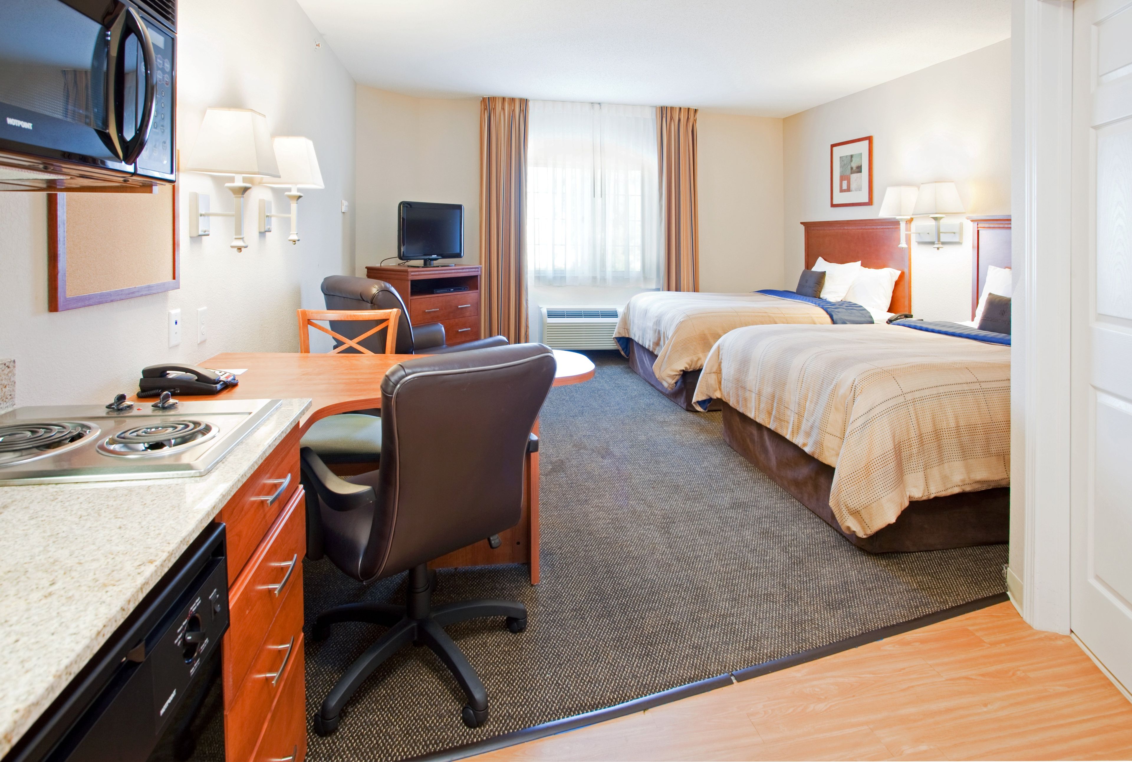 A hotel room with a kitchenette, a desk with a chair, two beds, a television, and a window with curtains.