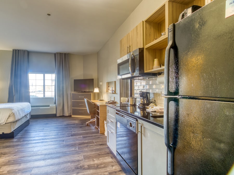 A compact hotel room with a kitchenette, a bed, a desk, and a window with curtains.