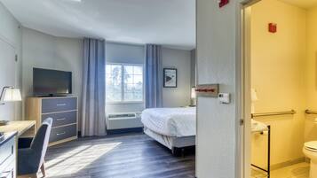 A hotel room with a bed, a desk with a chair, a television, a window with curtains, and a bathroom.