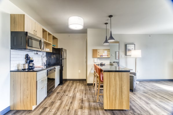 A modern kitchen with wooden cabinets, a microwave, and a refrigerator. There is a dining area with a table and chairs. - Candlewood Suites New Bern by IHG (New Bern)