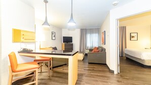 A modern hotel room with a wooden floor, a dining area with a table and chairs, a sofa, a bed, and a TV.
