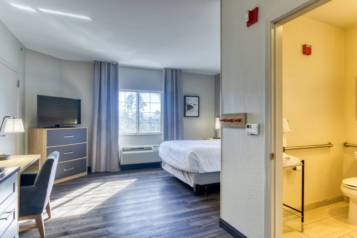 Une chambre d’hôtel comprenant un lit, un bureau avec une chaise, une télévision, une fenêtre avec des rideaux et une salle de bain avec des toilettes et un porte-serviettes.