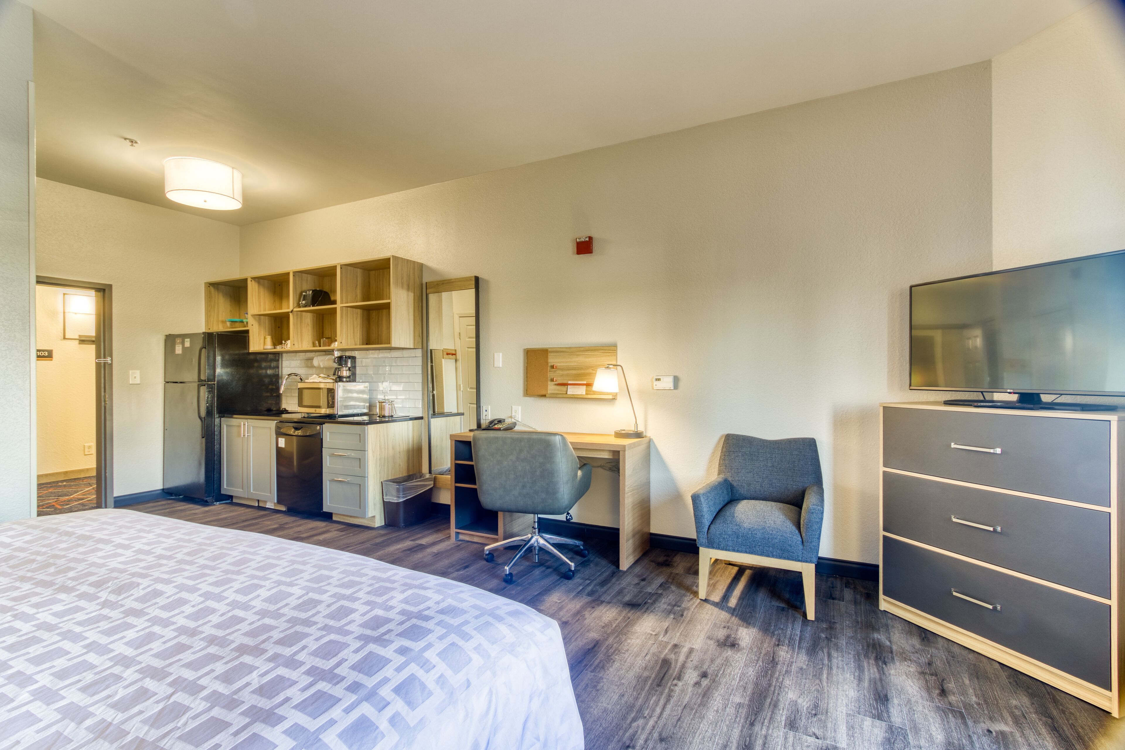 A hotel room with a bed, kitchenette, desk, two chairs, and a TV.