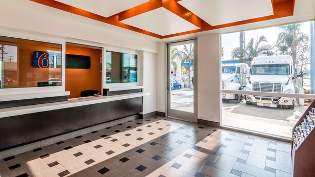 A modern reception area with a check-in counter, a digital display, and a large window offering a view of the street outside. - Motel 6 Bell Gardens, CA - Los Angeles (Bell Gardens)