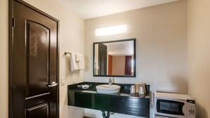 A bathroom with a dark wooden door, a white sink with a mirror above it, and a microwave on the countertop. - Motel 6 Bell Gardens, CA - Los Angeles (Bell Gardens)