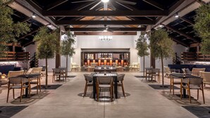 Un patio al aire libre con mesas y sillas, una zona de bar al fondo y árboles que dan sombra.