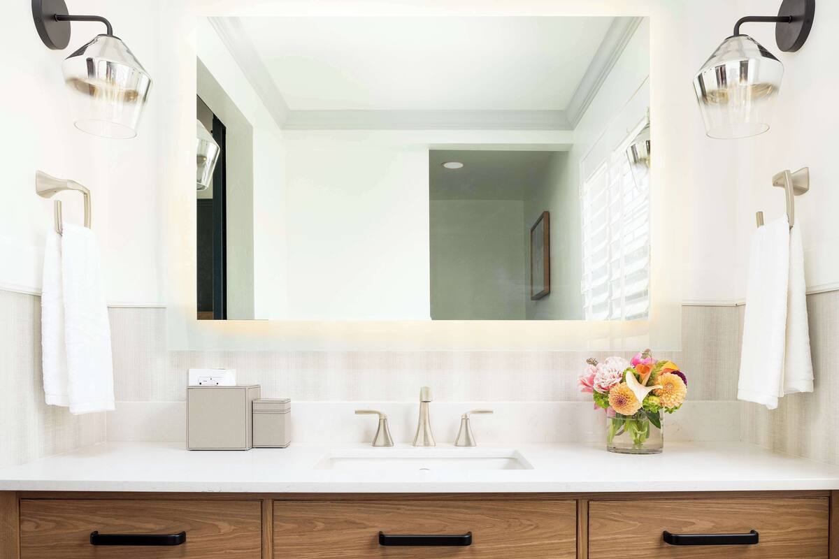 A modern bathroom with a large mirror, two wall-mounted lights, a white sink, and a vase of flowers.
