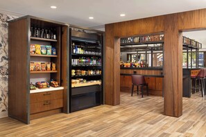 A modern bar area with wooden shelves stocked with snacks and beverages, a seating area with bar stools, and a view of the street through large windows.