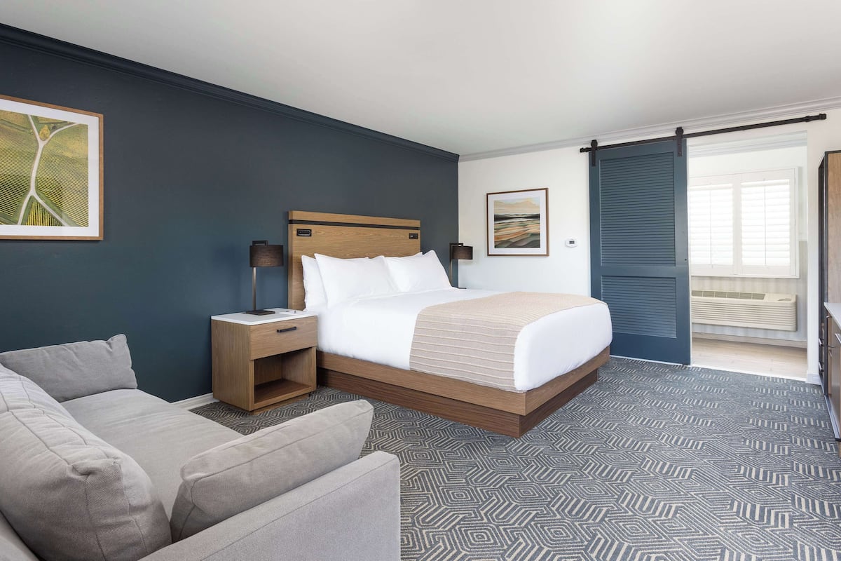A hotel room with a bed, a nightstand, a lamp, a sofa, and a large window with blinds.