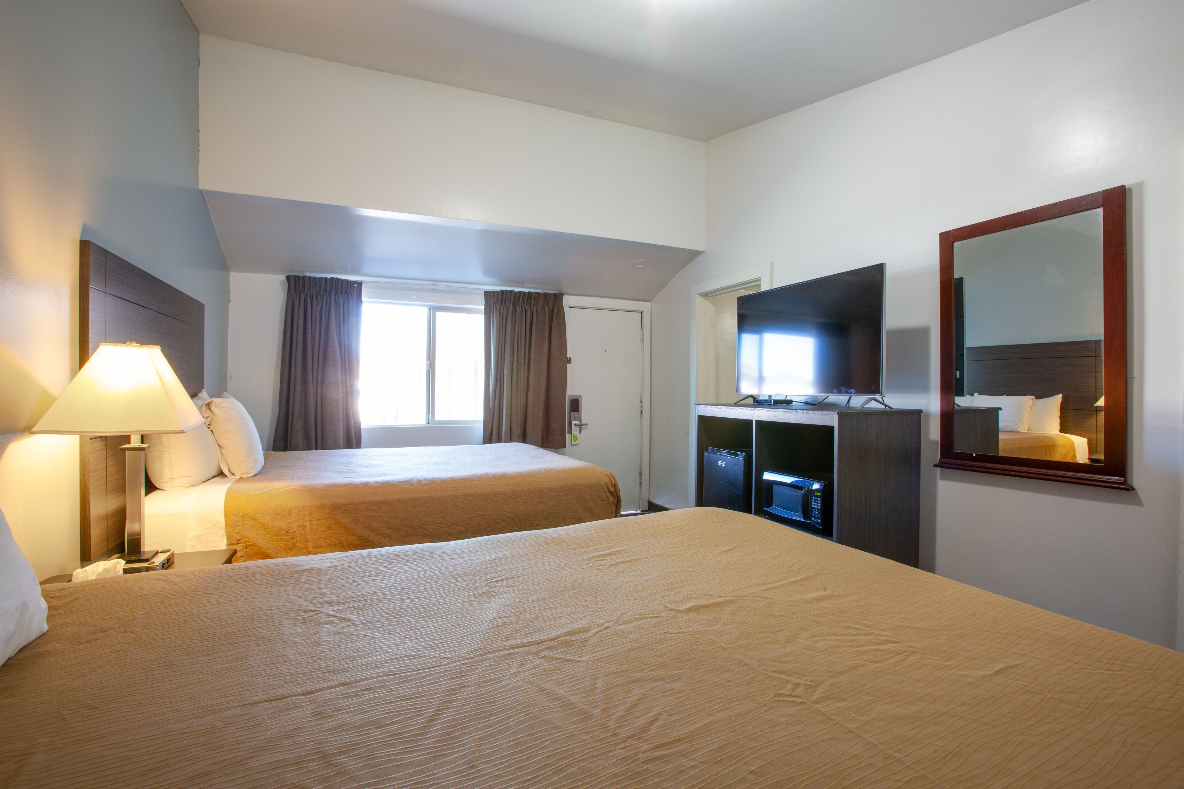 A hotel room with two beds, a desk with a computer, a mirror, and a window with curtains.