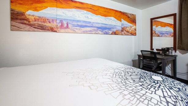 Habitación de hotel con un cuadro de paisaje grande, una cama con colcha de estampado geométrico, un escritorio con silla y un espejo en la pared.